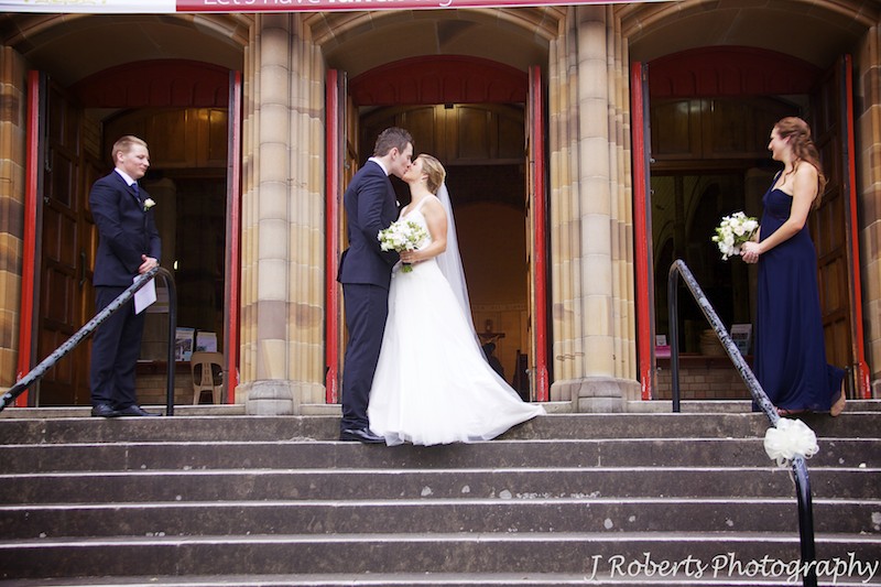 Bride and groom kissing outside church doors with attendants watching - wedding photography sydney
